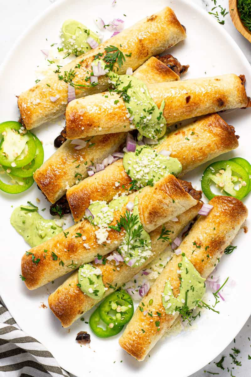 Large white platter with baked flautas garnished with fresh cilantro and avocado cream sauce