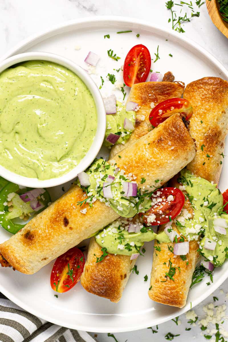 White plate with three baked flautas garnished with fresh cilantro and avocado cream sauce