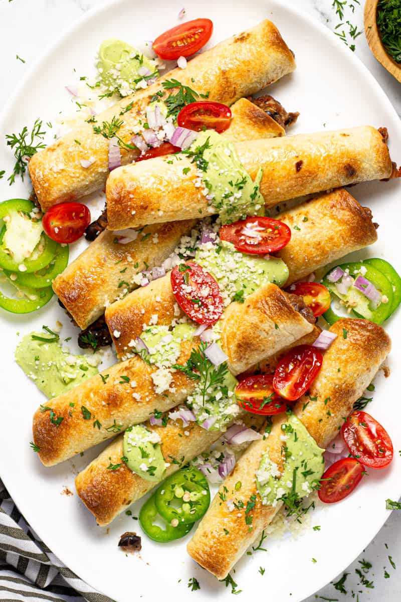 Large white platter with baked flautas garnished with fresh cilantro and avocado cream sauce
