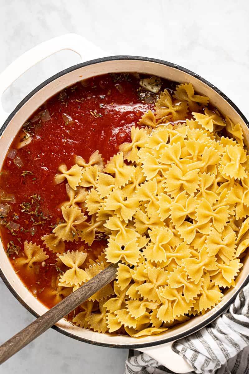 White Dutch oven with tomato sauce and uncooked bow tie noodles