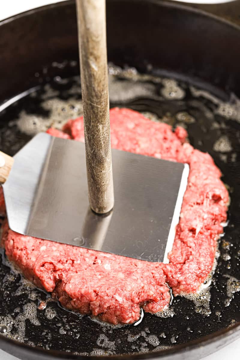A large spatula and wooden spoon smashing a ball of ground beef into a buttered cast iron skillet