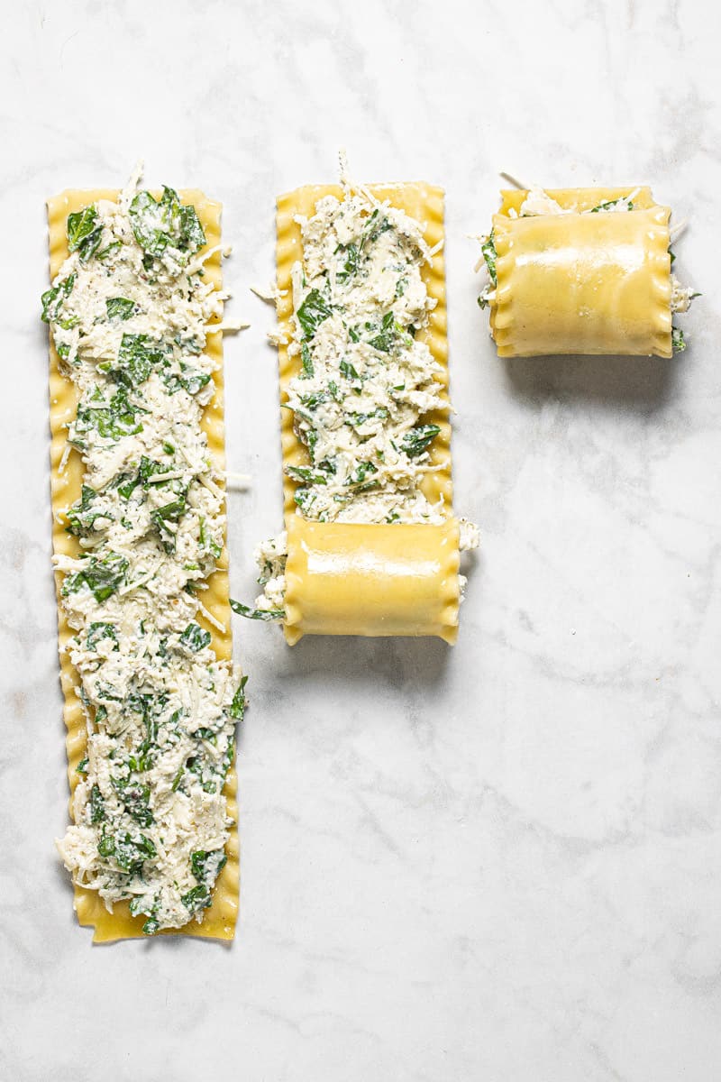 3 lasagna noodles with filling being rolled up on a white marble counter top