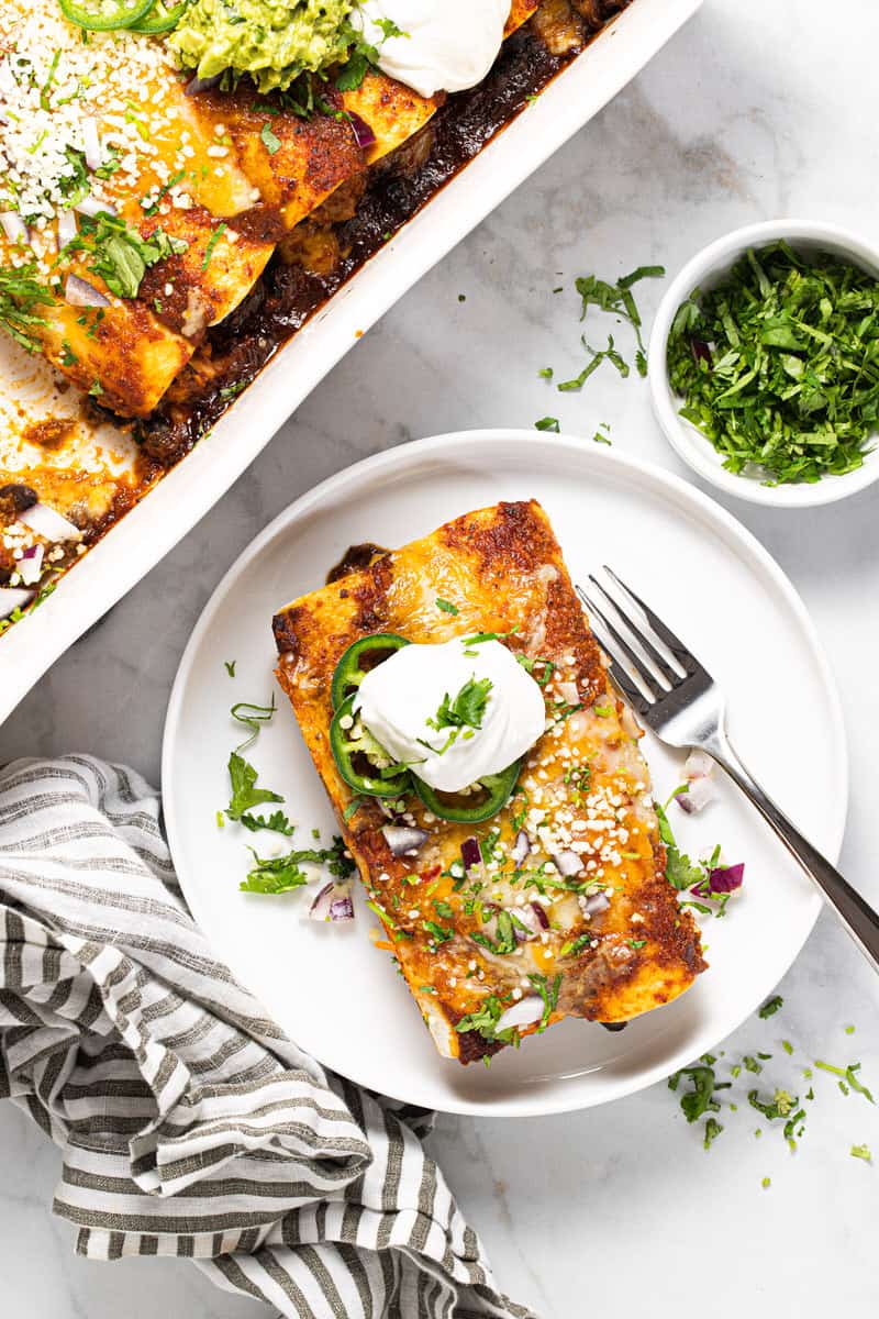 White plate with two cheesy chicken enchiladas topped with sour cream and cilantro