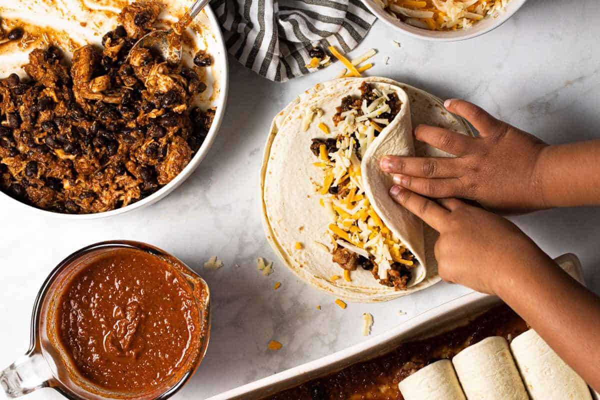 Two little hands rolling up a chicken enchilada filled with cheese