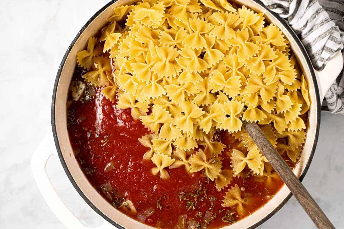White Dutch oven with tomato sauce and uncooked bow tie noodles