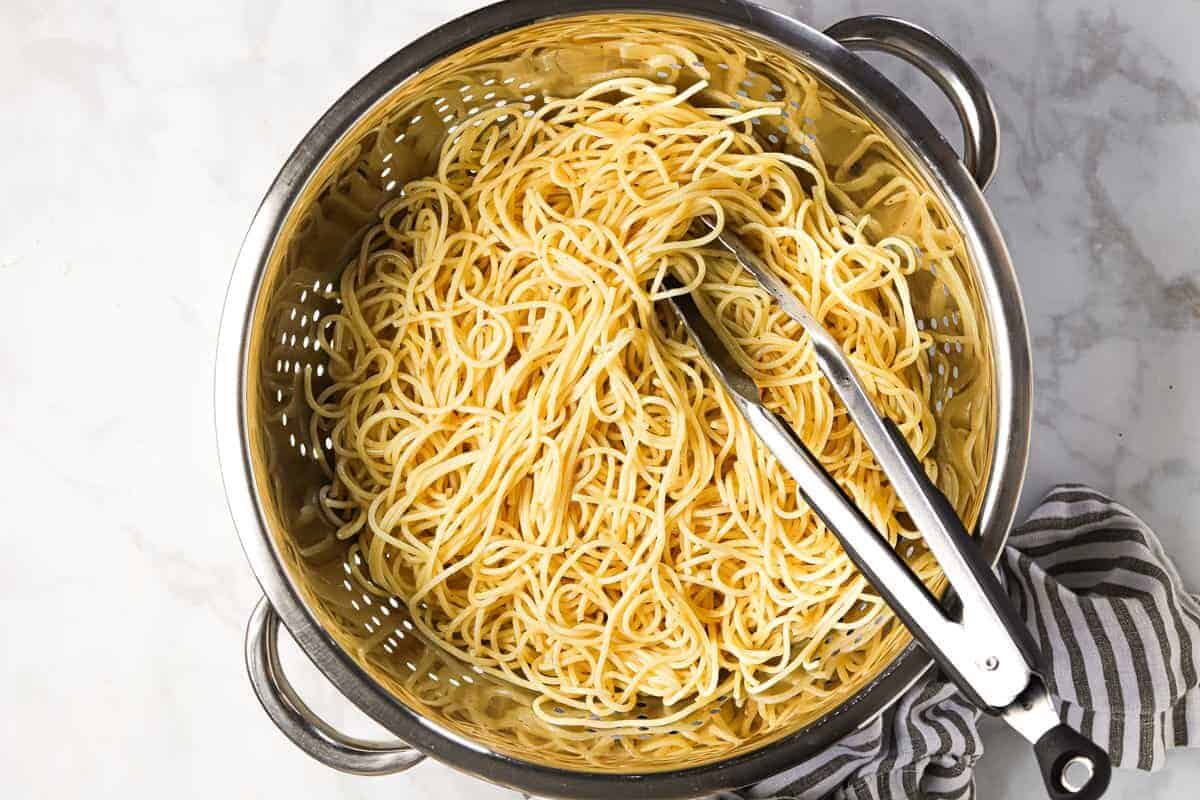 Cooked spaghetti in a metal strainer with metal tongs