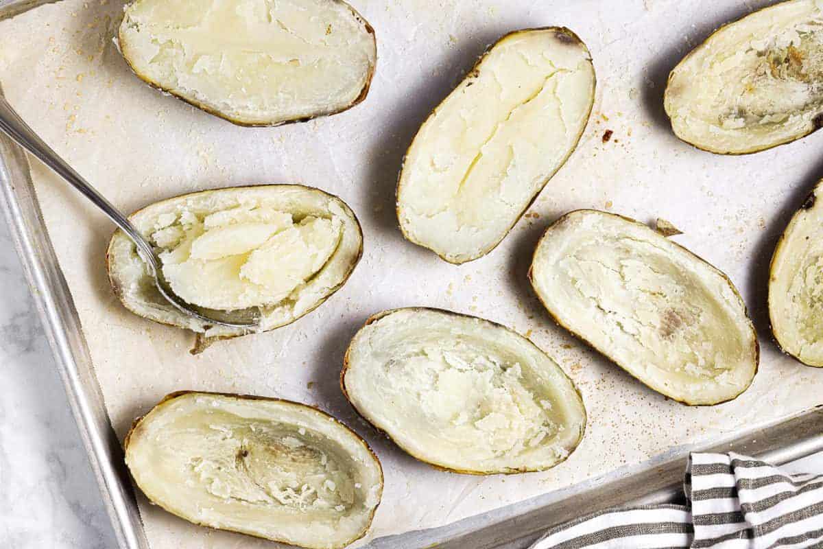 Baking sheet with halved baked potatoes with their centers being scooped out with a spoon 