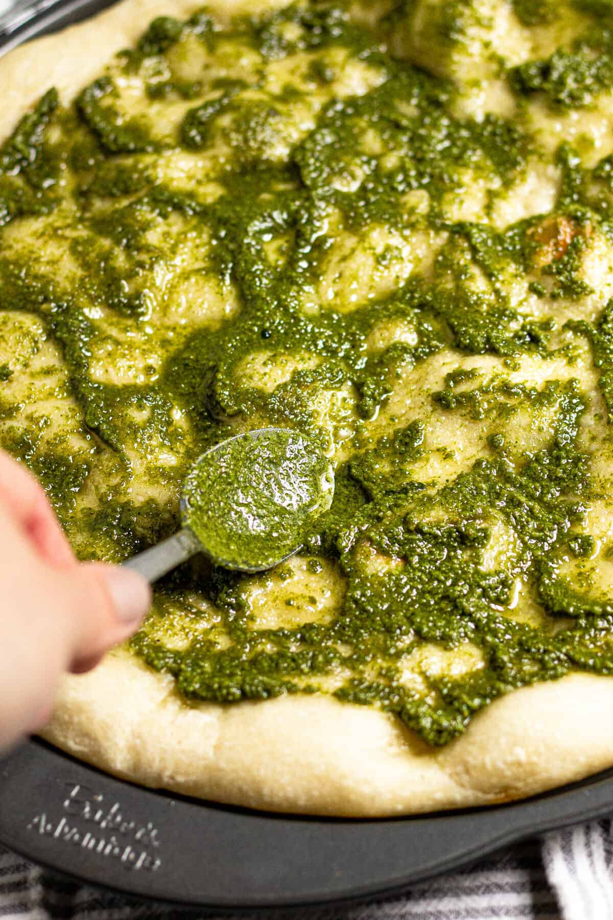 Basil pesto being spread across a pizza crust on a pizza pan 