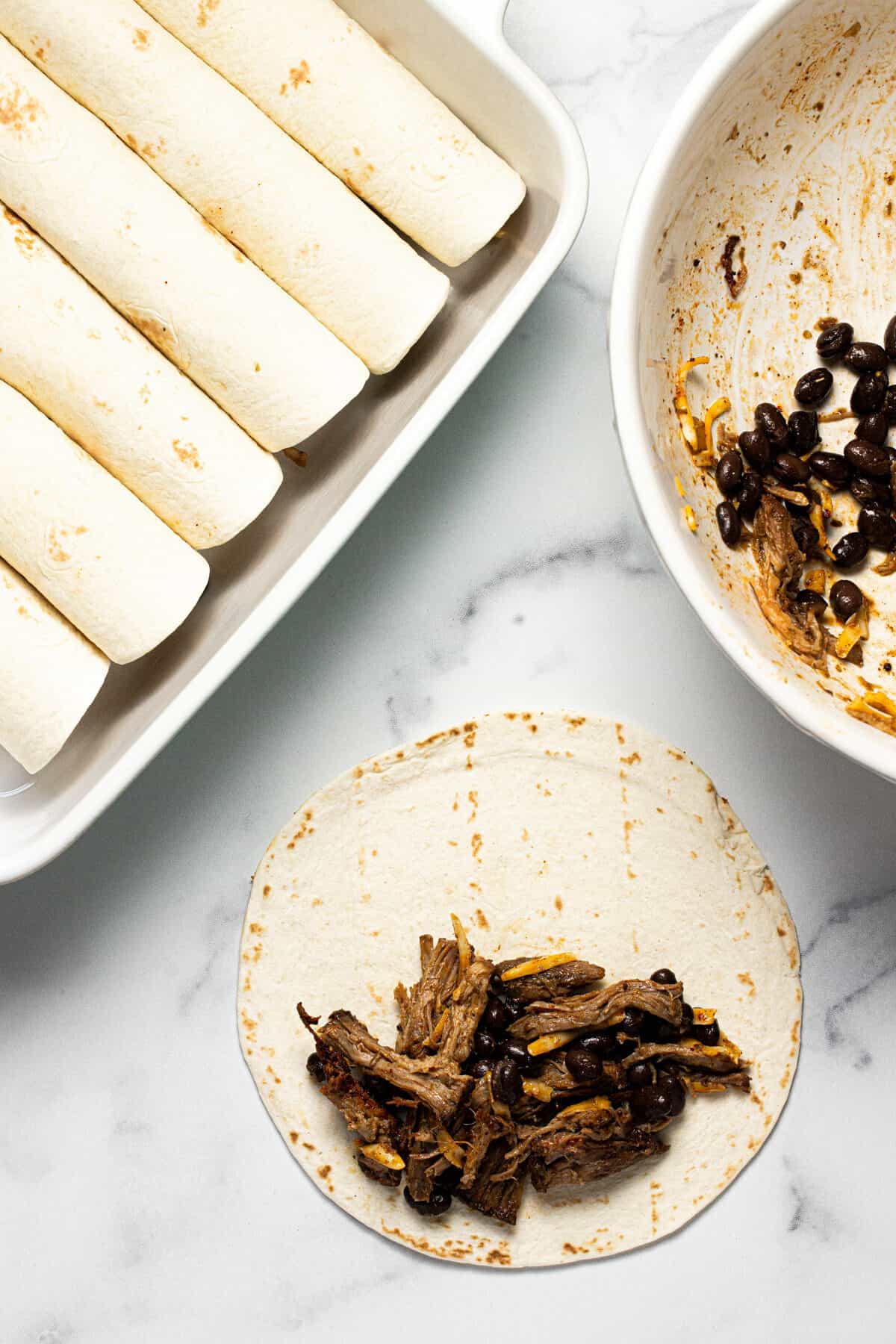 Pork filling in the middle of a flour tortilla next to pan of rolled enchiladas