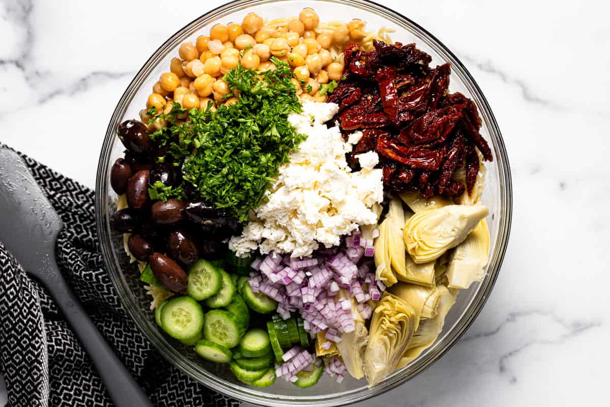 Glass bowl filled with ingredients to make Mediterranean pasta salad