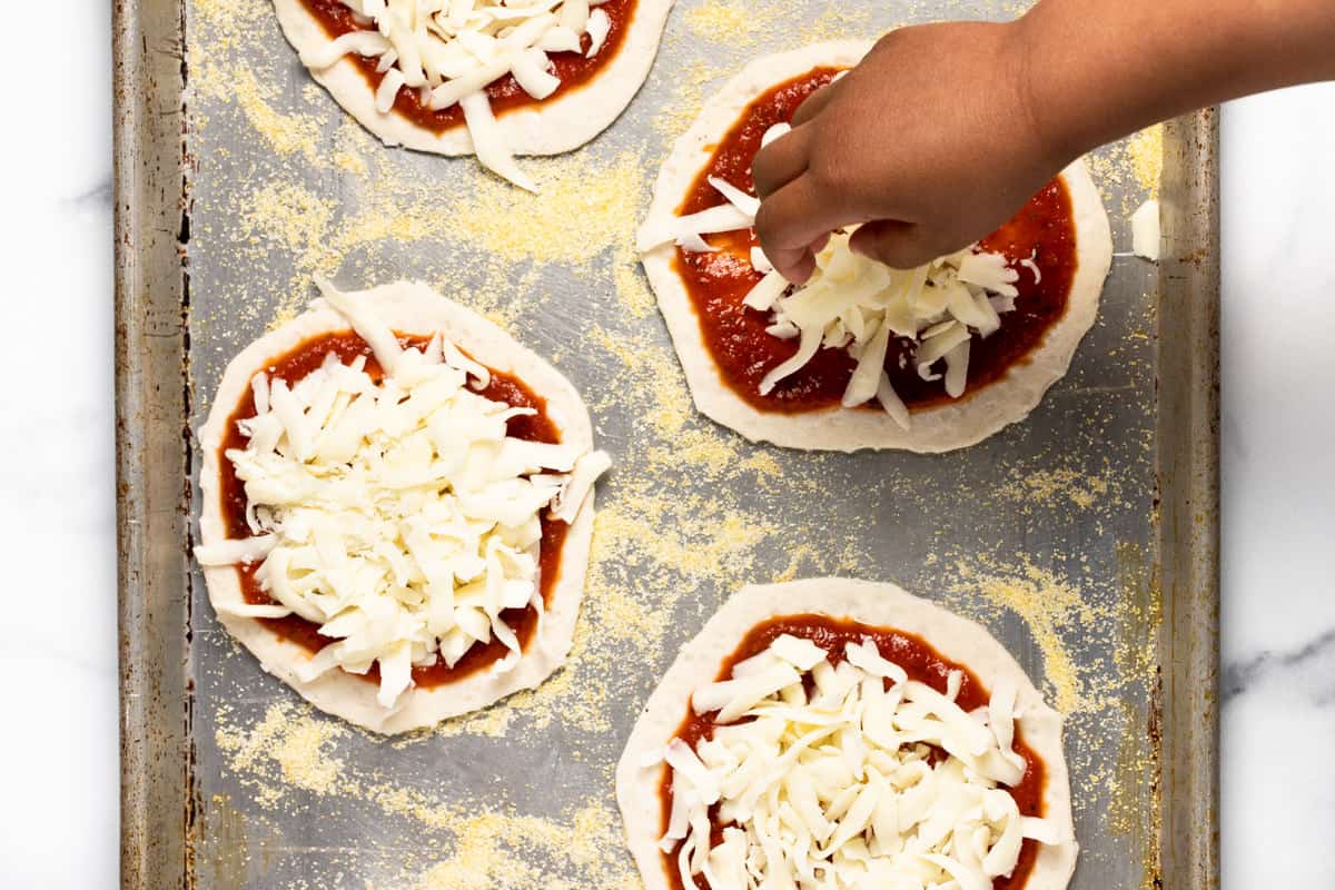 A small hand adding a handful of cheese to mini pizzas