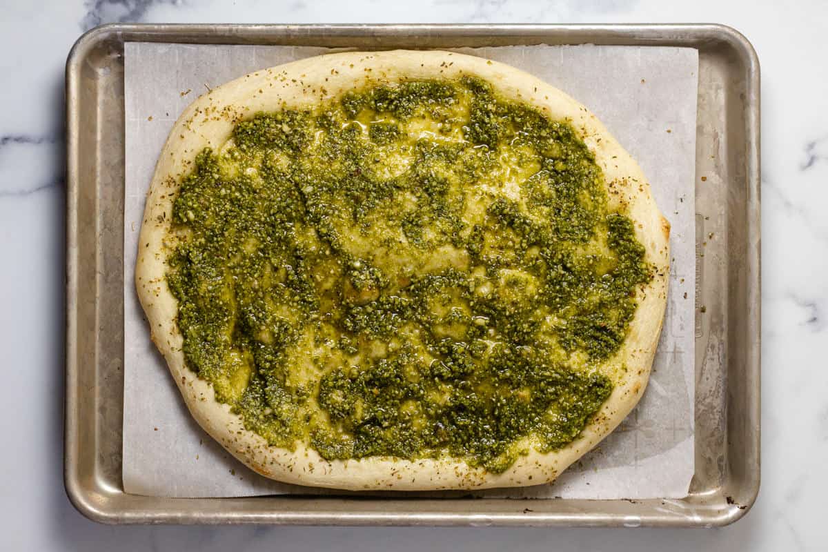 Parchment lined baked sheet with par-baked pizza crust topped with pesto