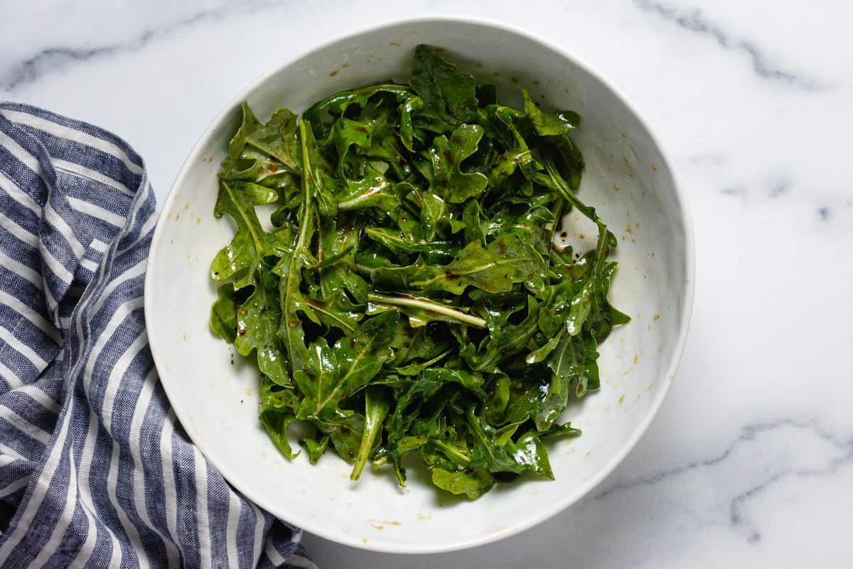 Arugula in a large white bowl tossed with balsamic vinegar and olive oil