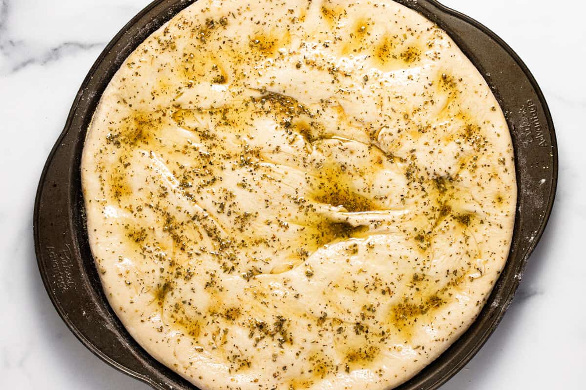 Pizza crust on a pizza pan with a layer of olive oil and herbs