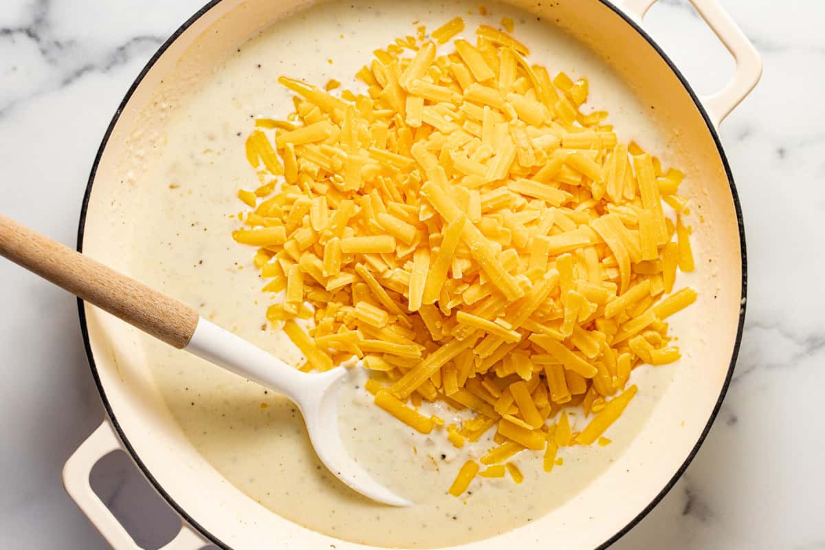 Cream sauce in a large white pan with shredded sharp cheddar