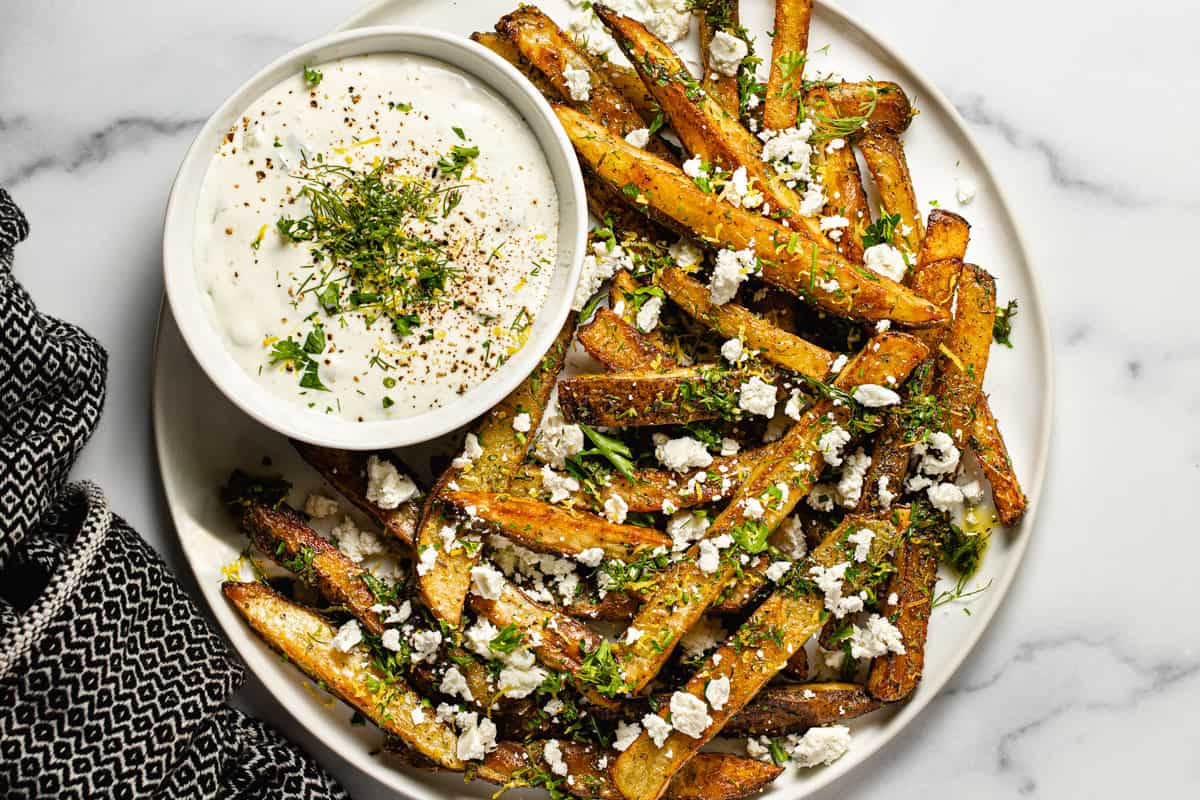 Large white plate filled with homemade Greek fries and a bowl of dill yogurt sauce