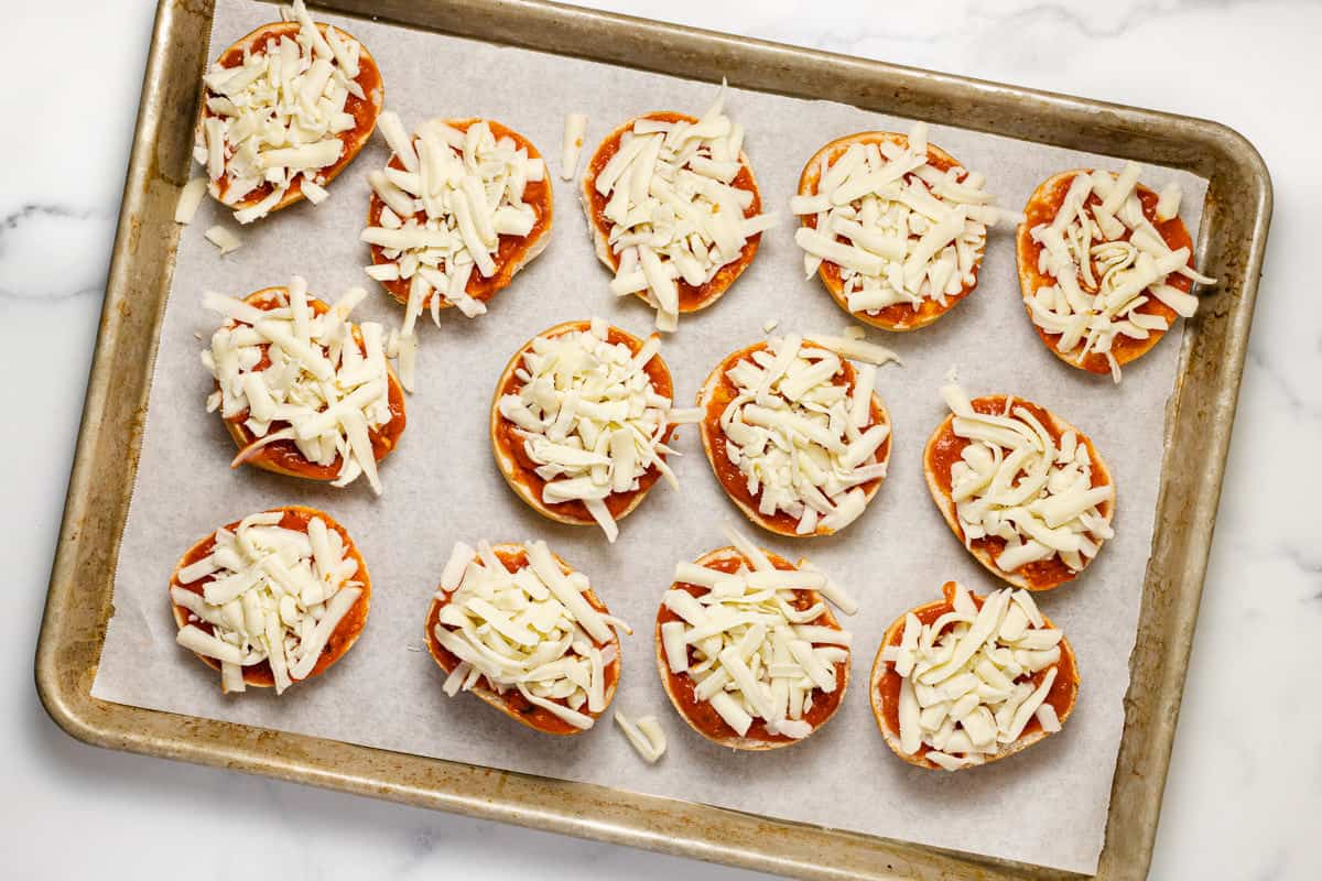 Plain mini bagels topped with pizza sauce and cheese on a parchment lined baking sheet