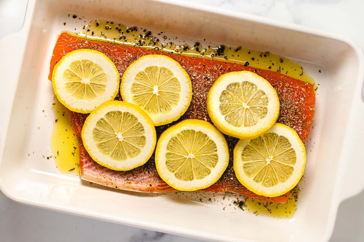 White baking dish with a large seasoned salmon filet topped with sliced lemons.