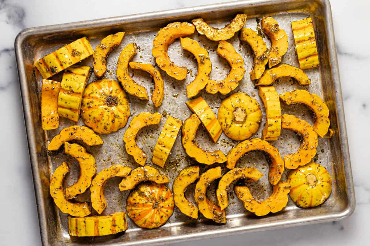 Sliced delicata squash tossed in oil and herbs on a small baking sheet
