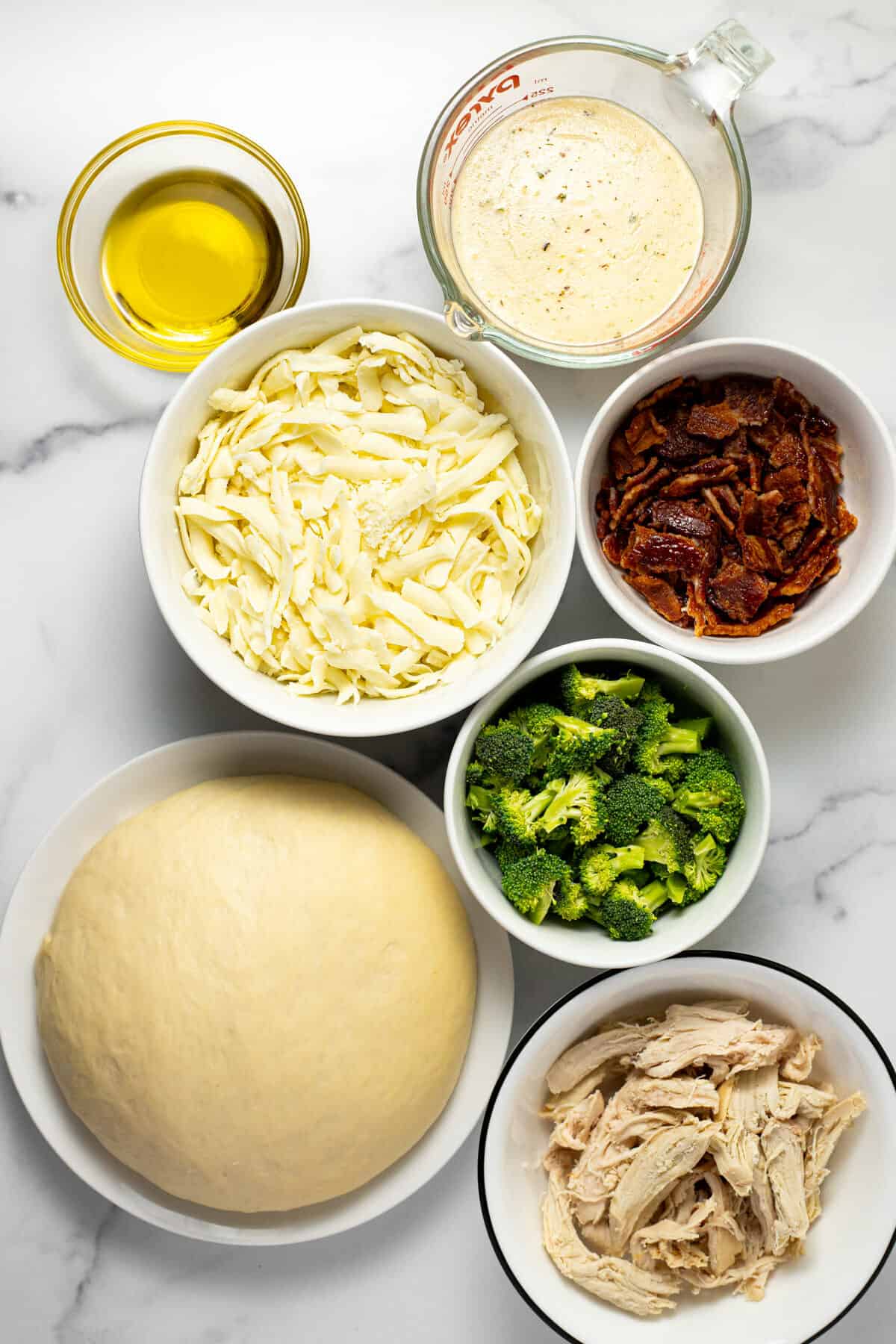 Marble counter top with bowls of ingredients to make chicken alfredo pizza