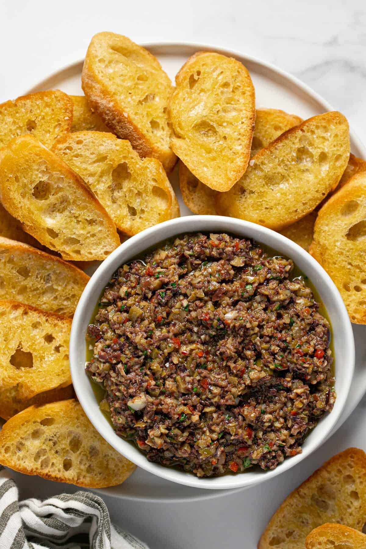 White bowl filled with homemade olive tapenade on a plate filled with crostini