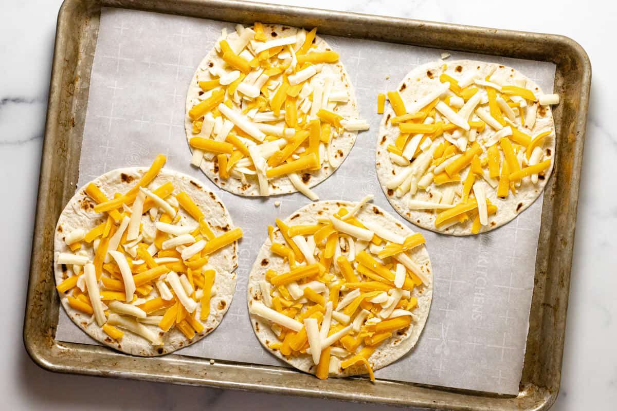 Four flour tortillas topped with cheese on a parchment lined baking sheet