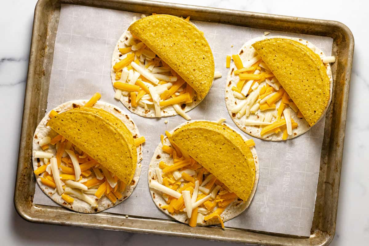 Four flour tortillas topped with cheese and a crispy taco shell on a parchment lined baking sheet