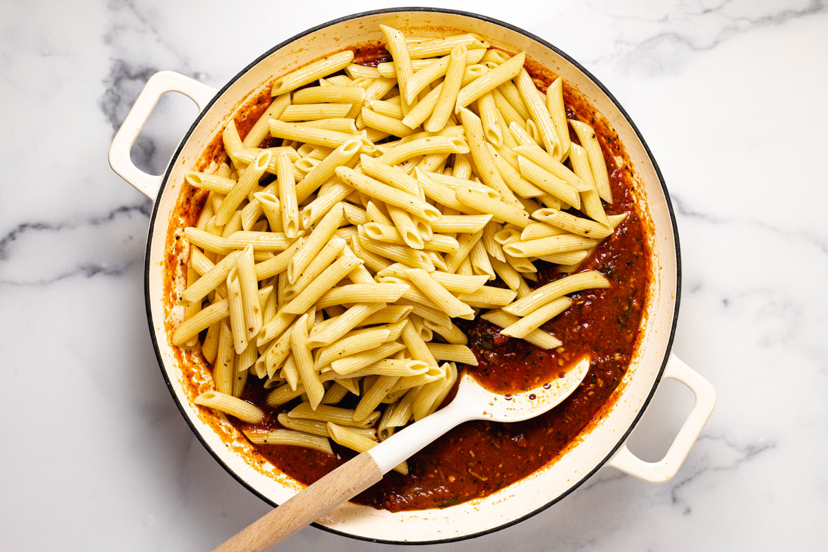 Penne pasta being added to a large pot of Pomodoro sauce.