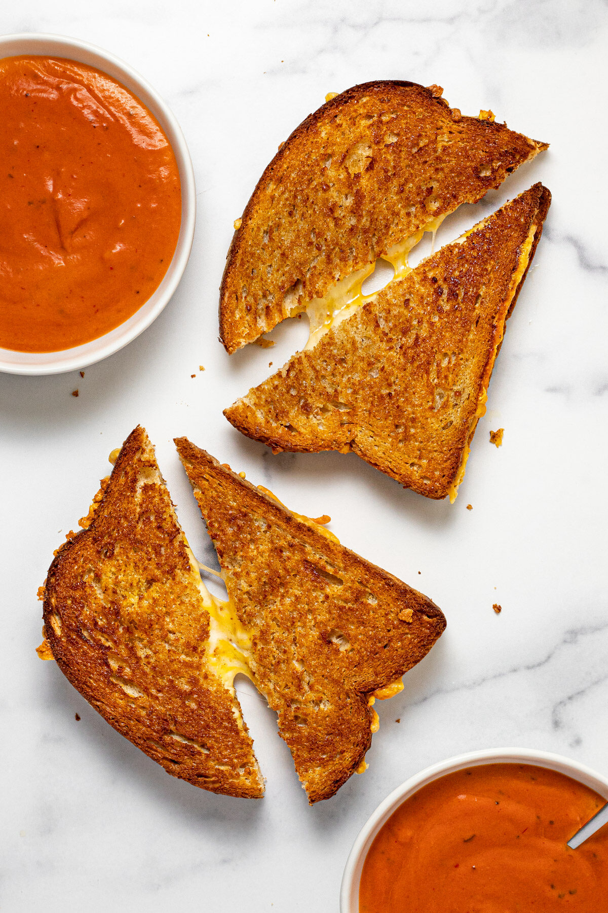 Two grilled cheese sandwiches sliced in half with melty cheese oozing out with two bowls of tomato soup