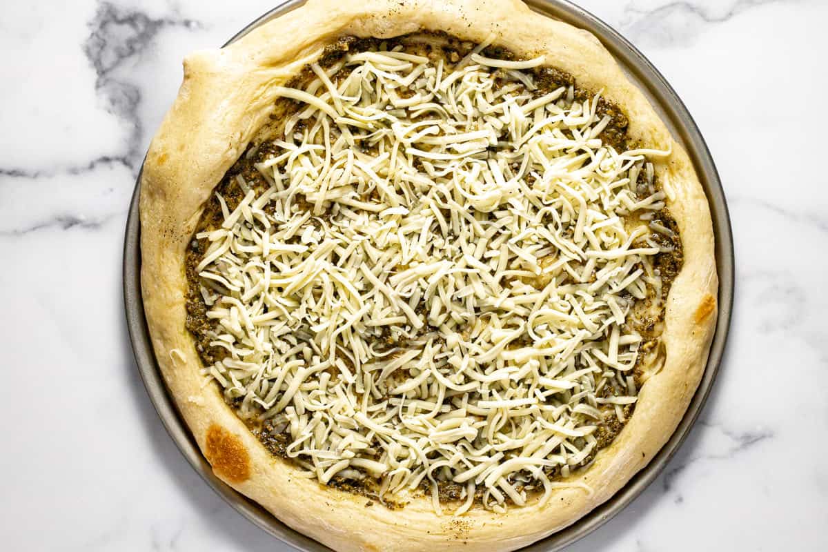 Pizza crust on a pizza pan topped with basil pesto and shredded cheese