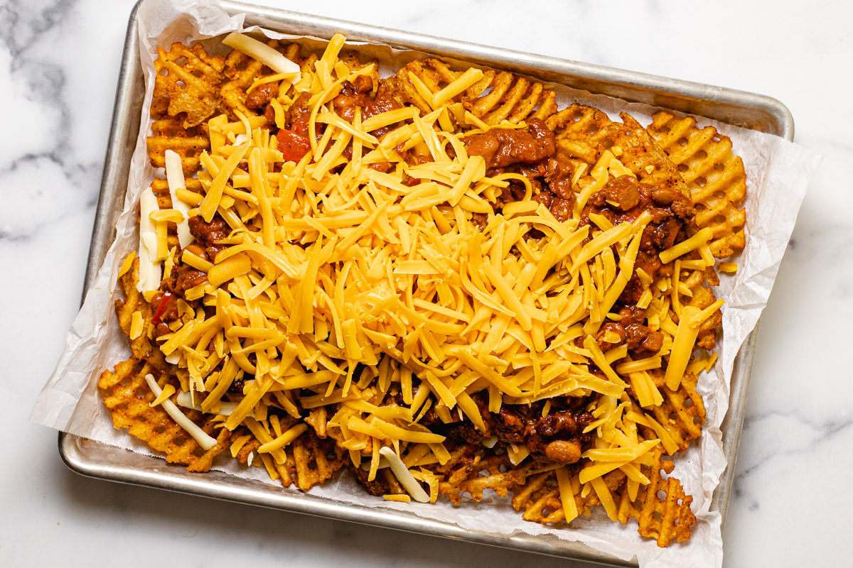 Crispy waffle fries on a parchment lined baking sheet topped with shredded cheese and chili