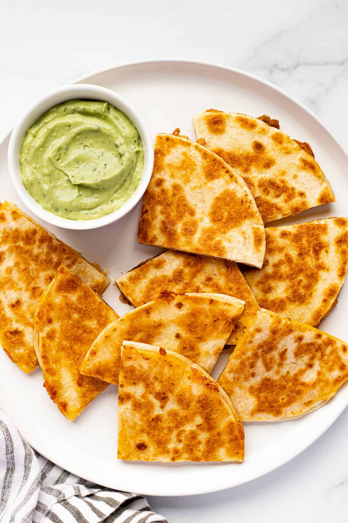 White plate filled with sliced quesadillas and a bowl of creamy avocado sauce