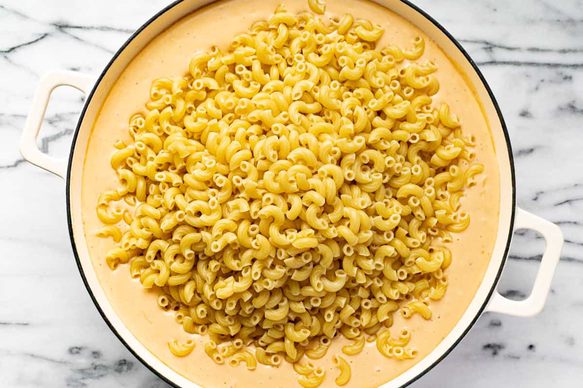 Macaroni noodles being added to a creamy cheese sauce in a white pan