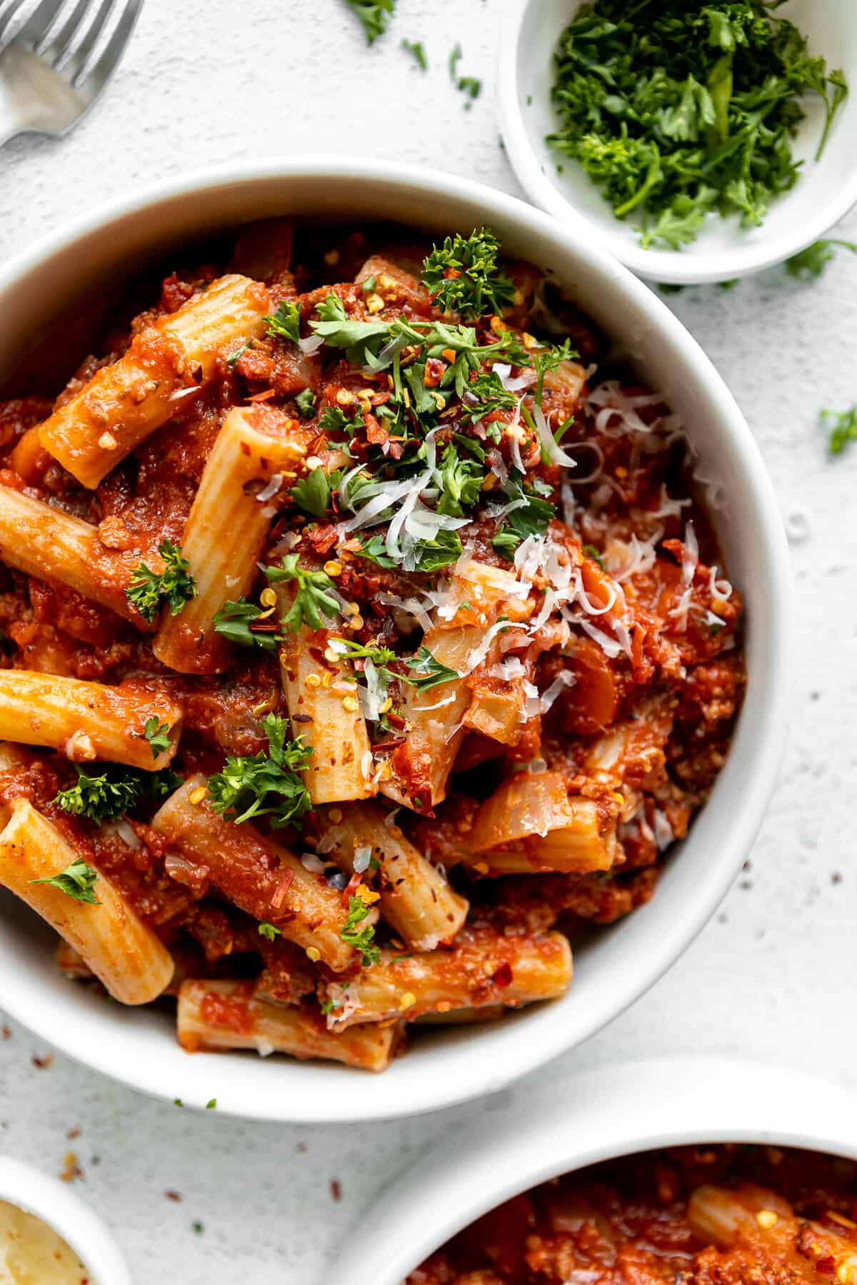White bowl filled with rigatoni bolognese garnished with Parmesa and parsley.