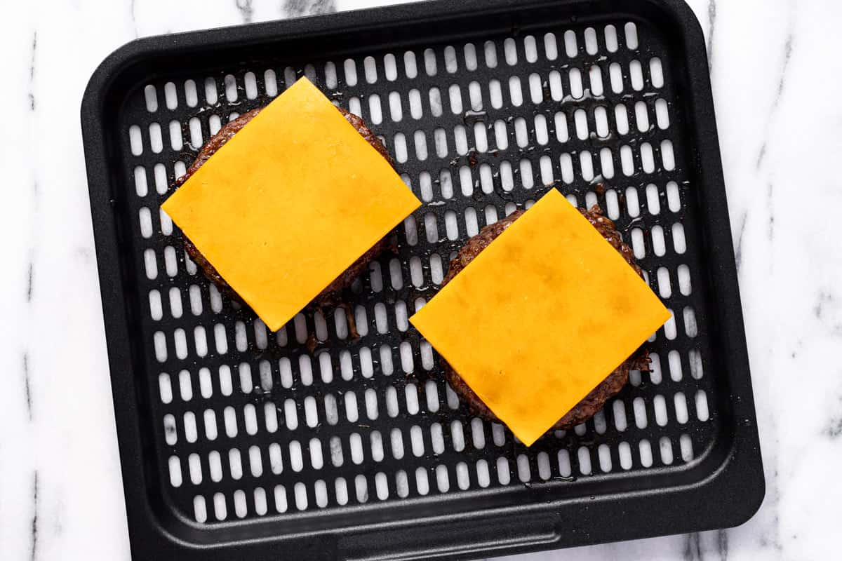 Two slices of cheese on top of hamburgers on an air fryer tray.