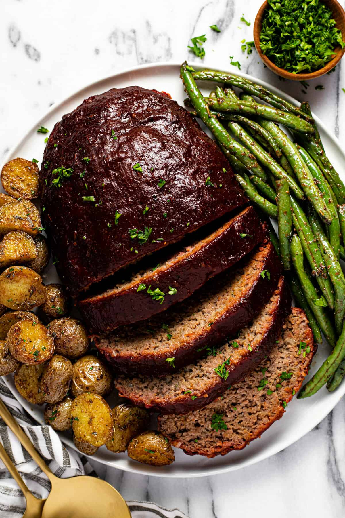 Sliced meatloaf on a white plate with roasted potatoes and green beans.