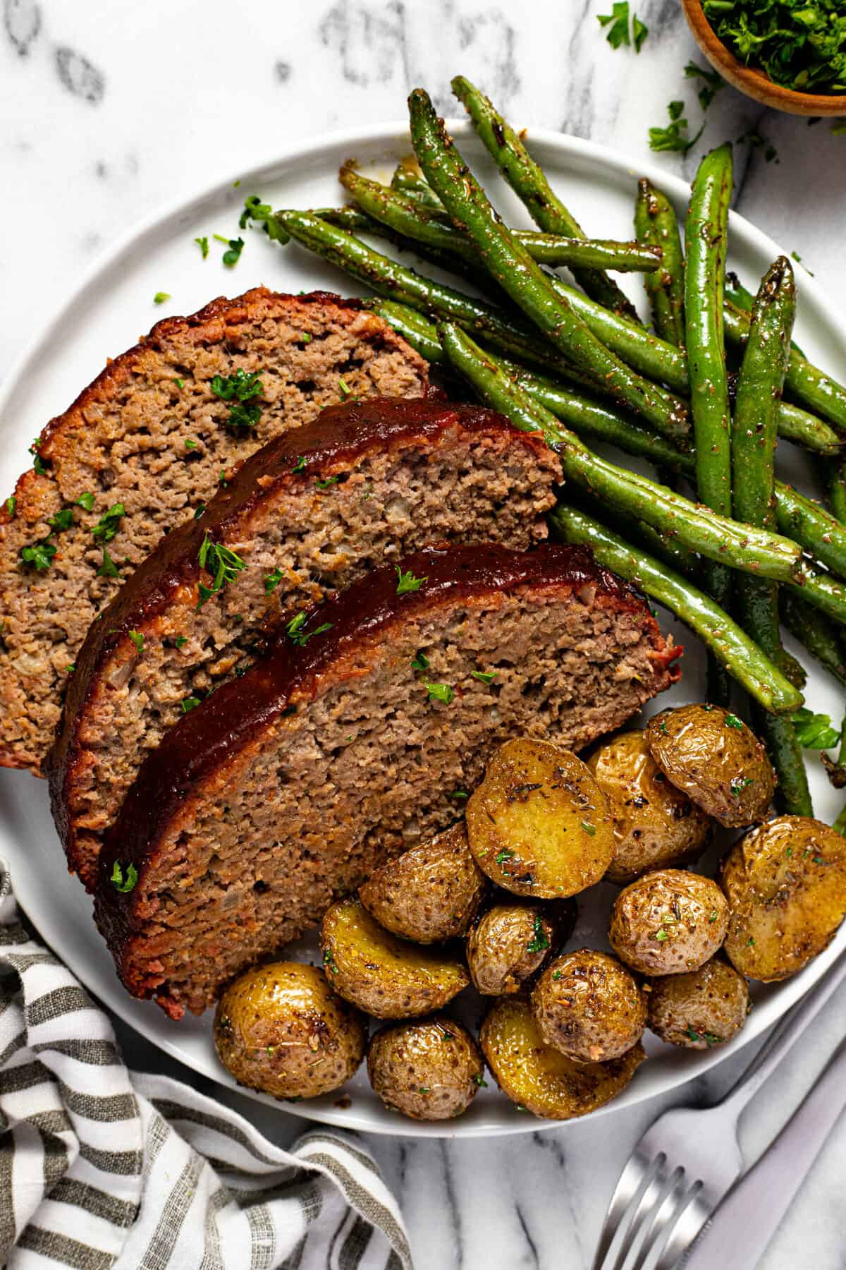White plate with sliced meatloaf roasted potatoes and green beans.