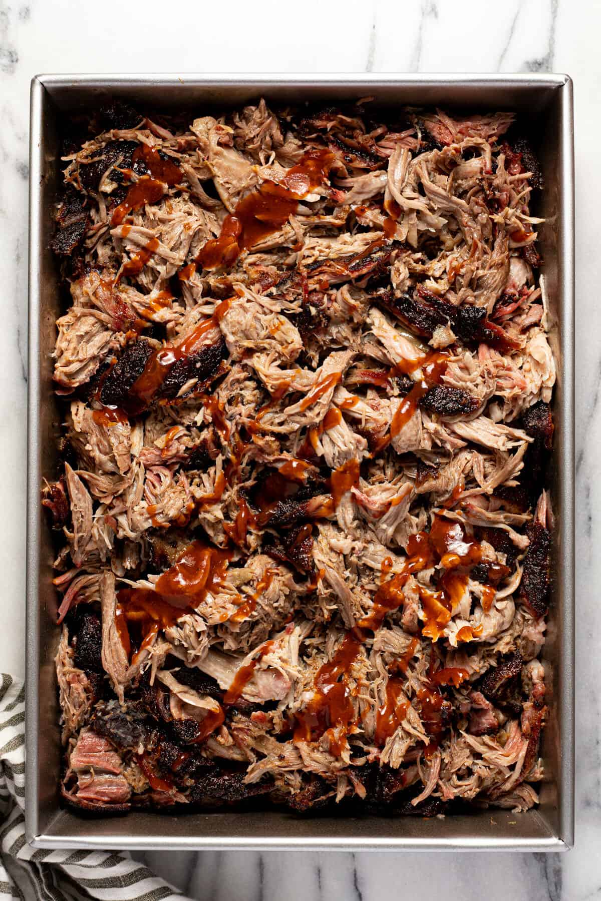 Large metal pan of shredded pork butt drizzled with BBQ sauce