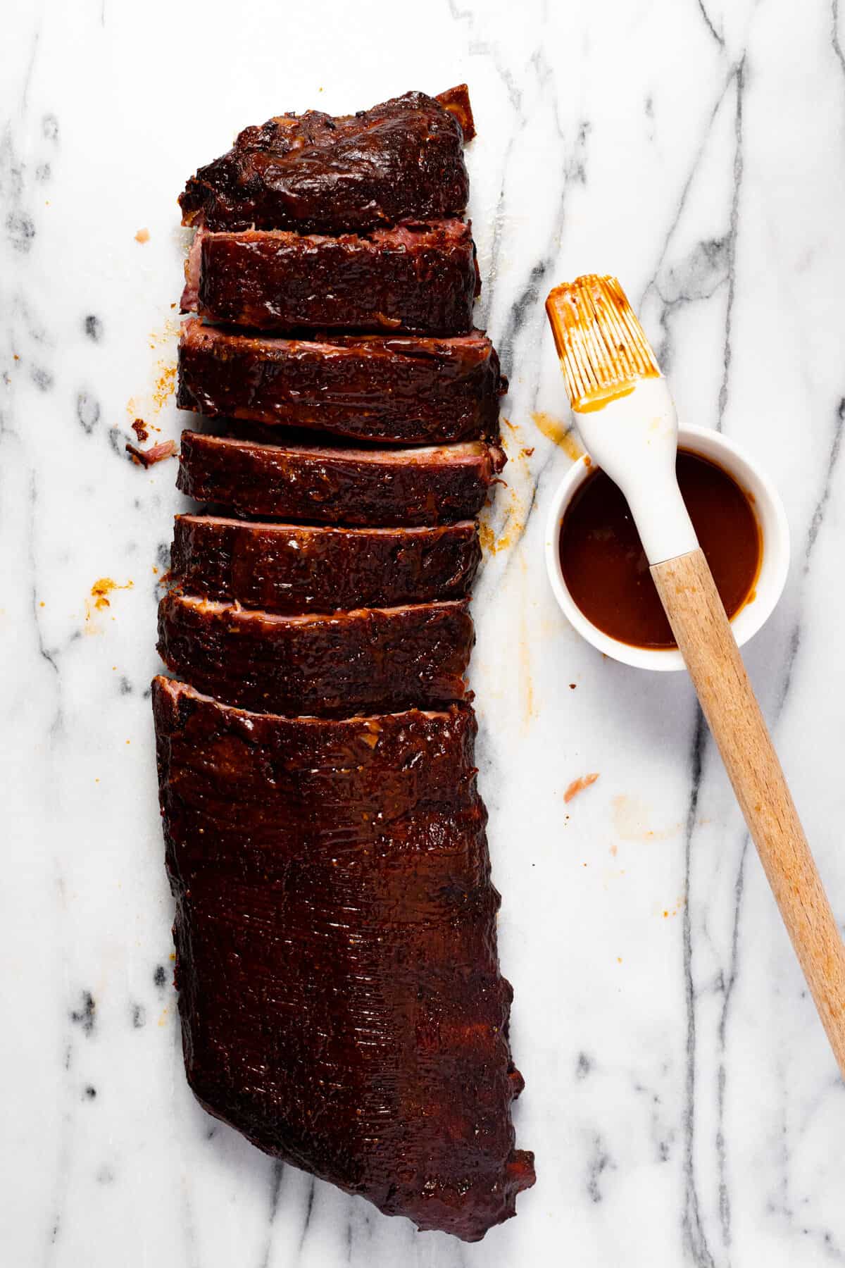 White marble counter top with a rack of ribs, half of them sliced, and a bowl of BBQ sauce.