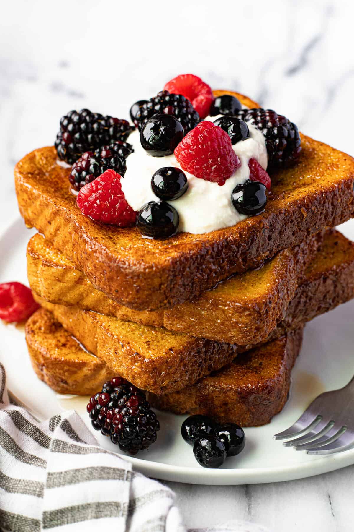 White plate stacked with 4 slices of French toasted topped with berries and cream.