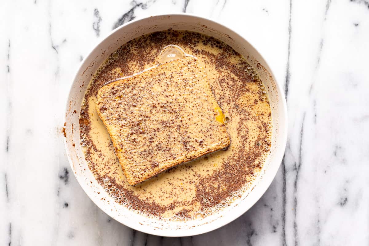 Thick slice of bread dipping in French toast batter in a white bowl.