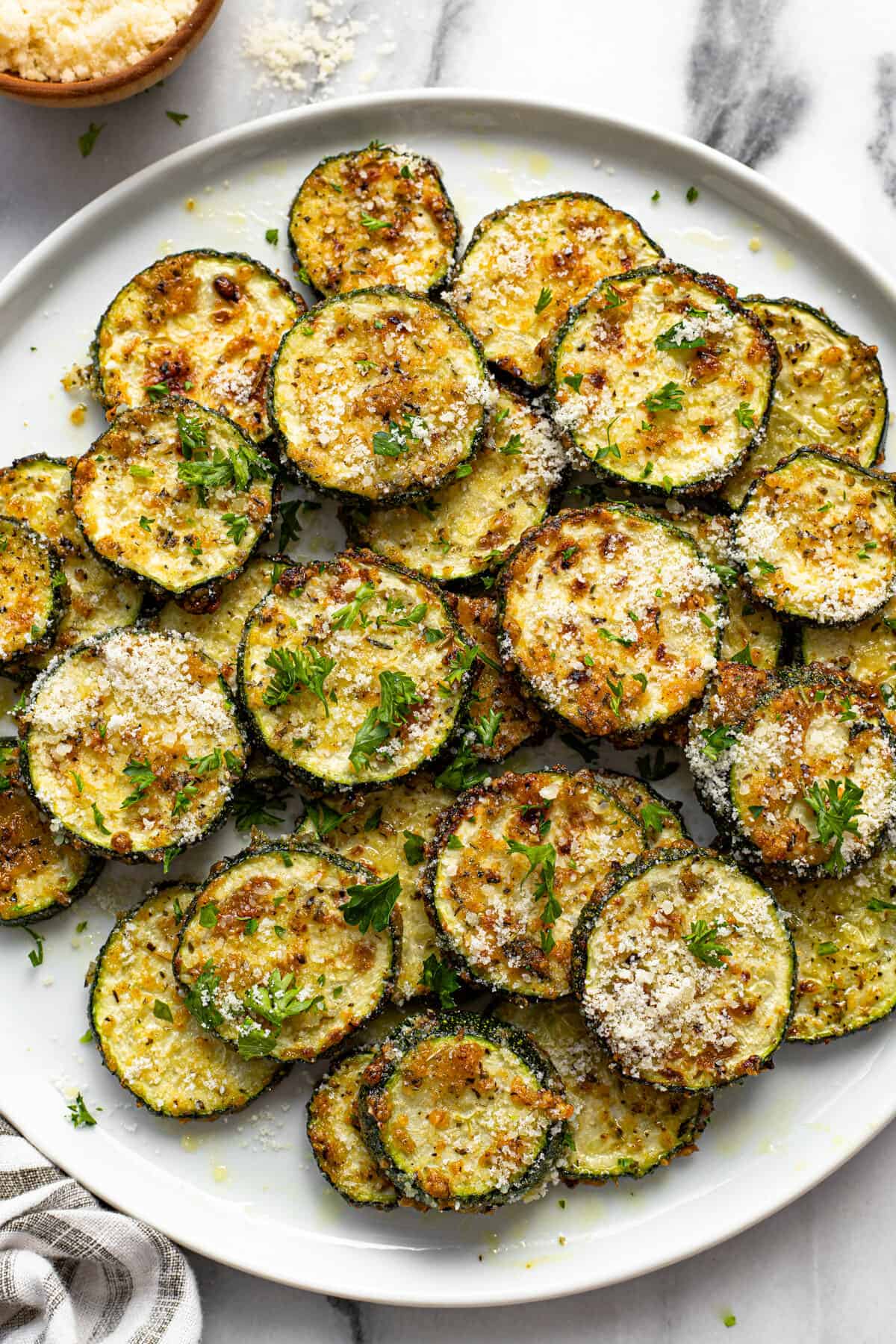 White plate filled with sliced air fryer zucchini garnished with parsley and Parmesan cheese.
