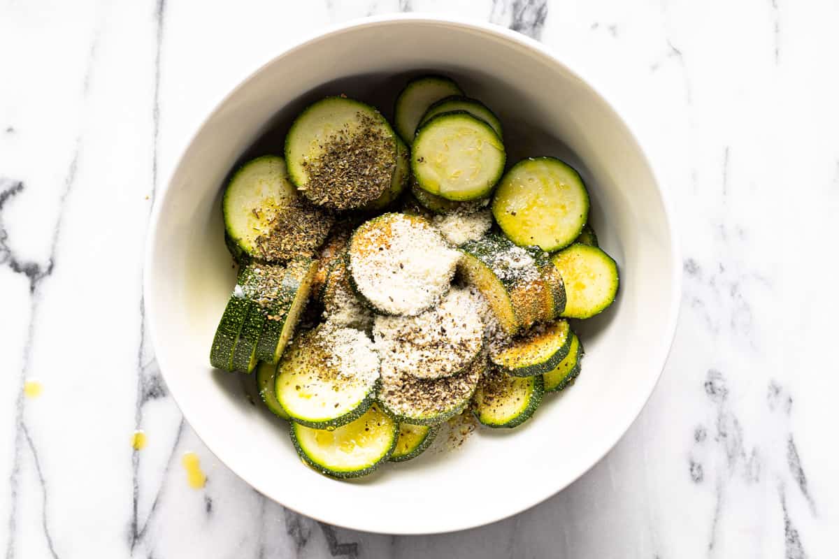 White bowl filled with sliced zucchini, olive oil, Parmesan cheese, herbs, and spices.
