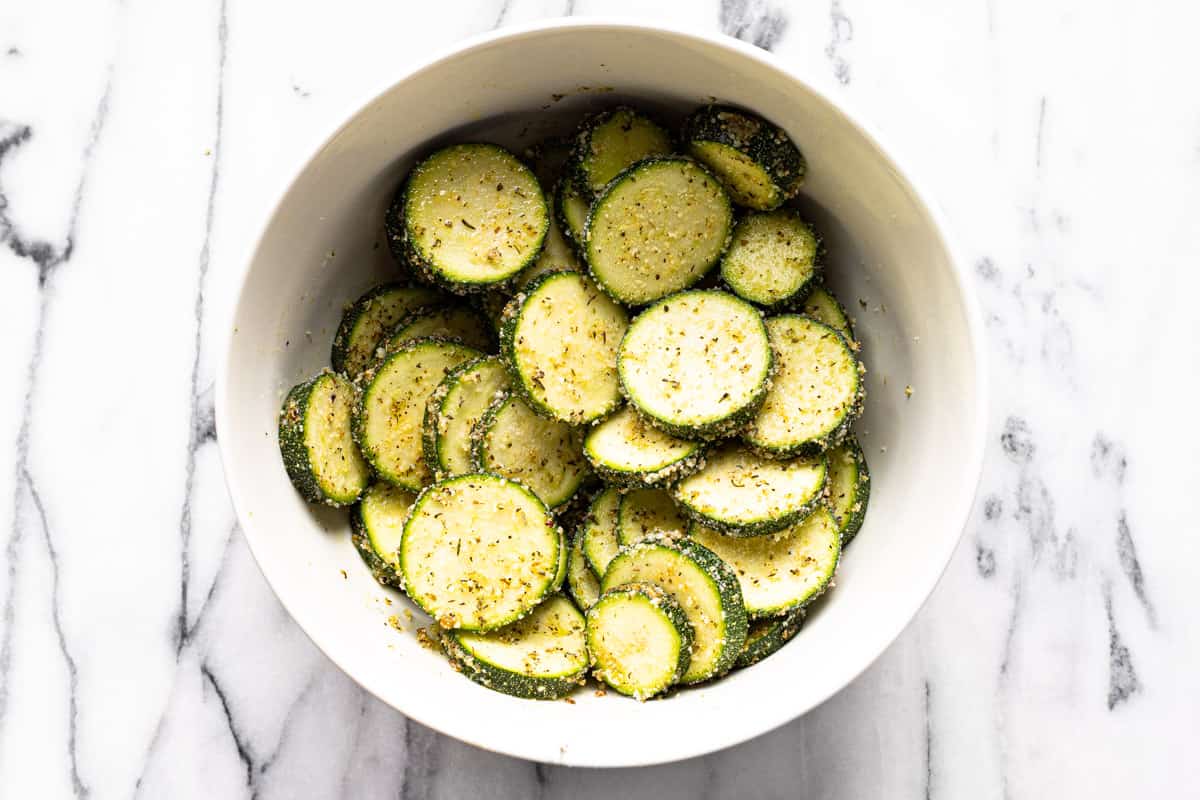 White bowl filled with sliced zucchini tossed in olive oil, Parmesan cheese, herbs, and spices.