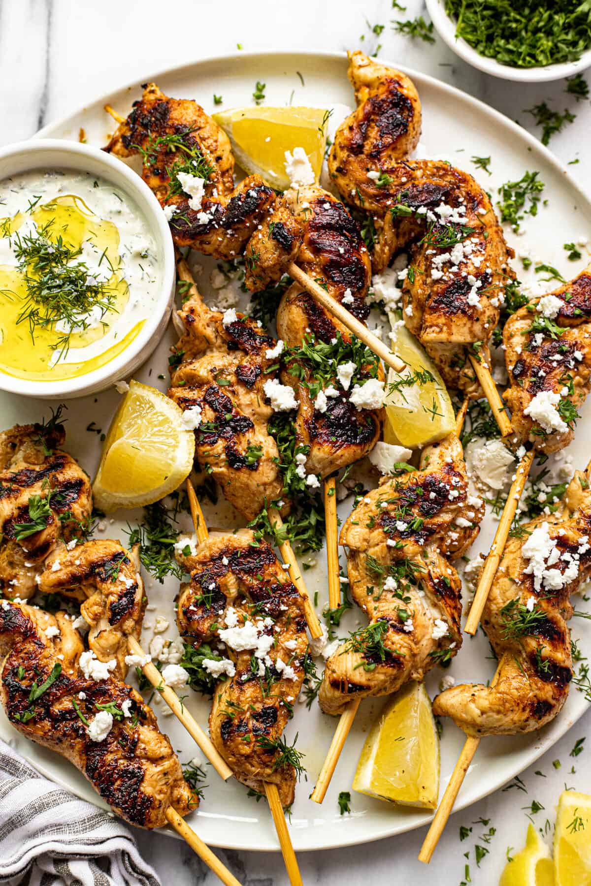 White plate filled with Greek chicken kebabs garnished with parsley, dill, and crumbled feta.