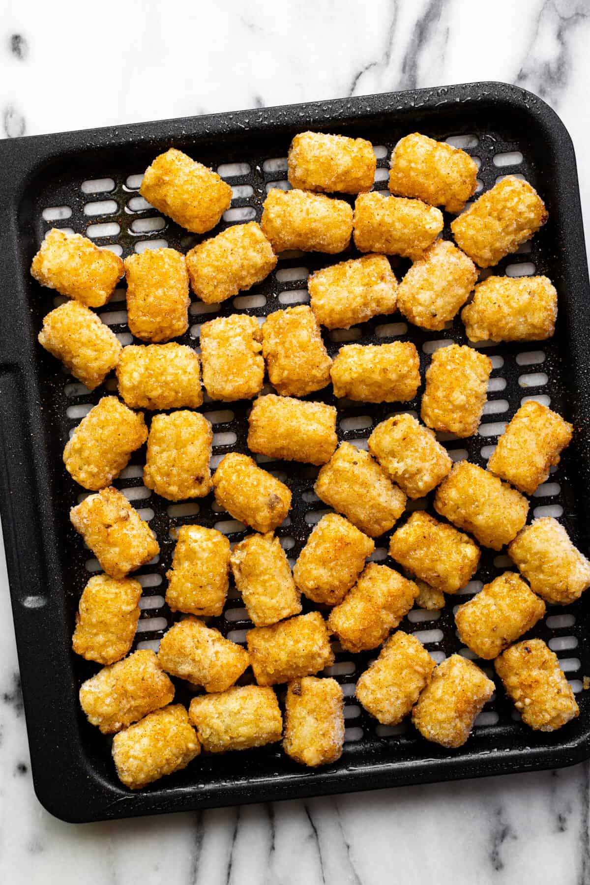 Black air fryer tray with frozen tater tots on it.