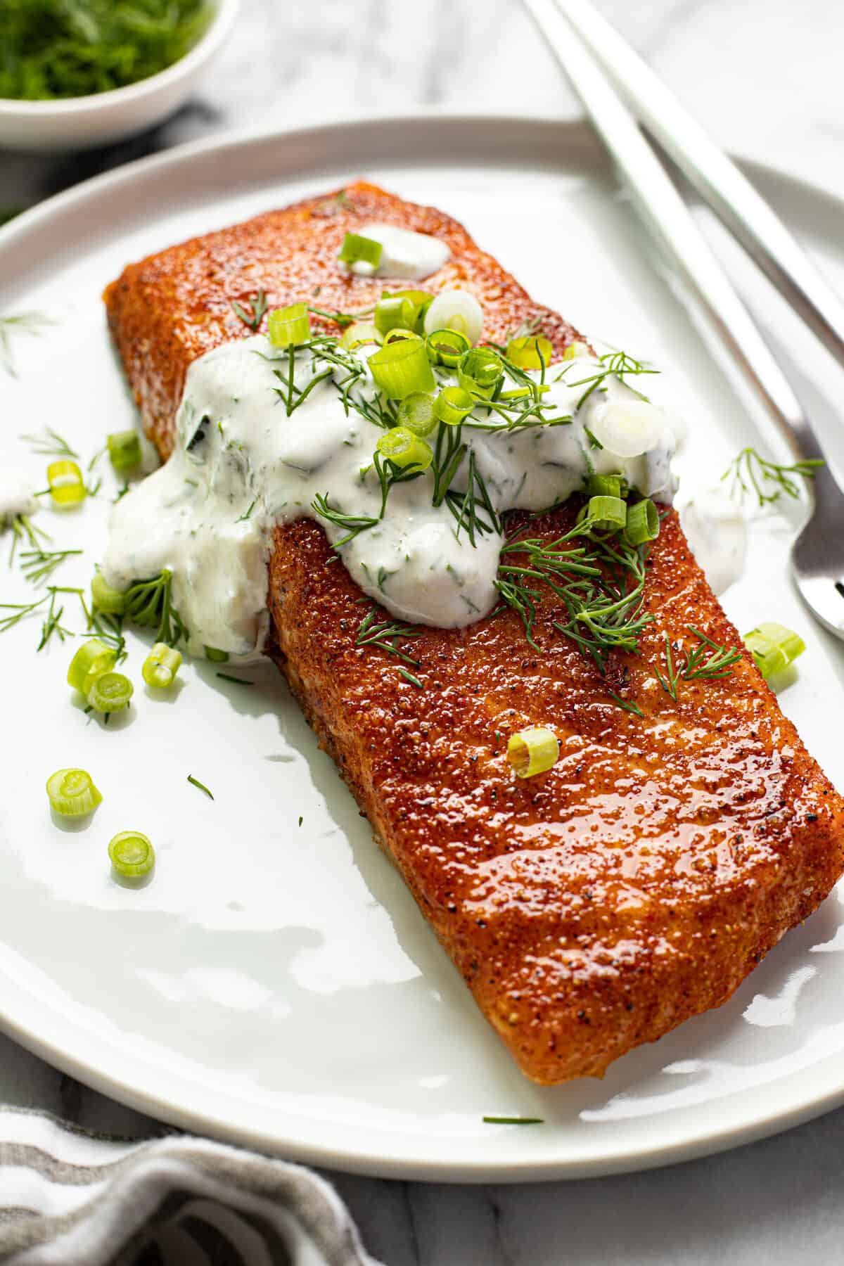 Close up shot of smoked salmon on a white plate garnished with dill sauce and sliced green onions.