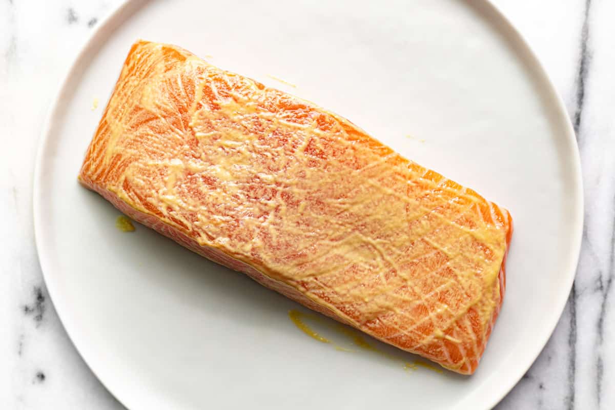 A salmon fillet rubbed with Dijon mustard on a white plate.