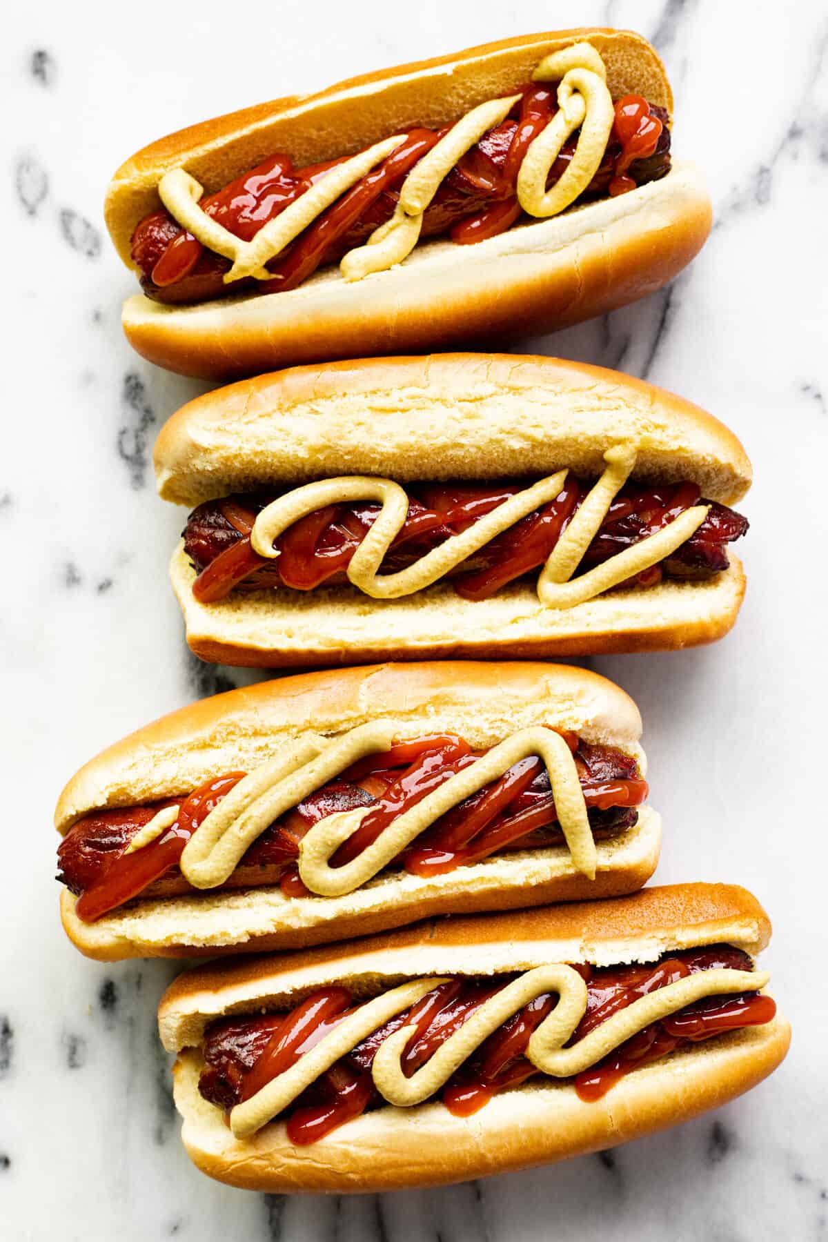 4 air fried hot dogs in buns topped with ketchup and mustard on a white marble counter top.