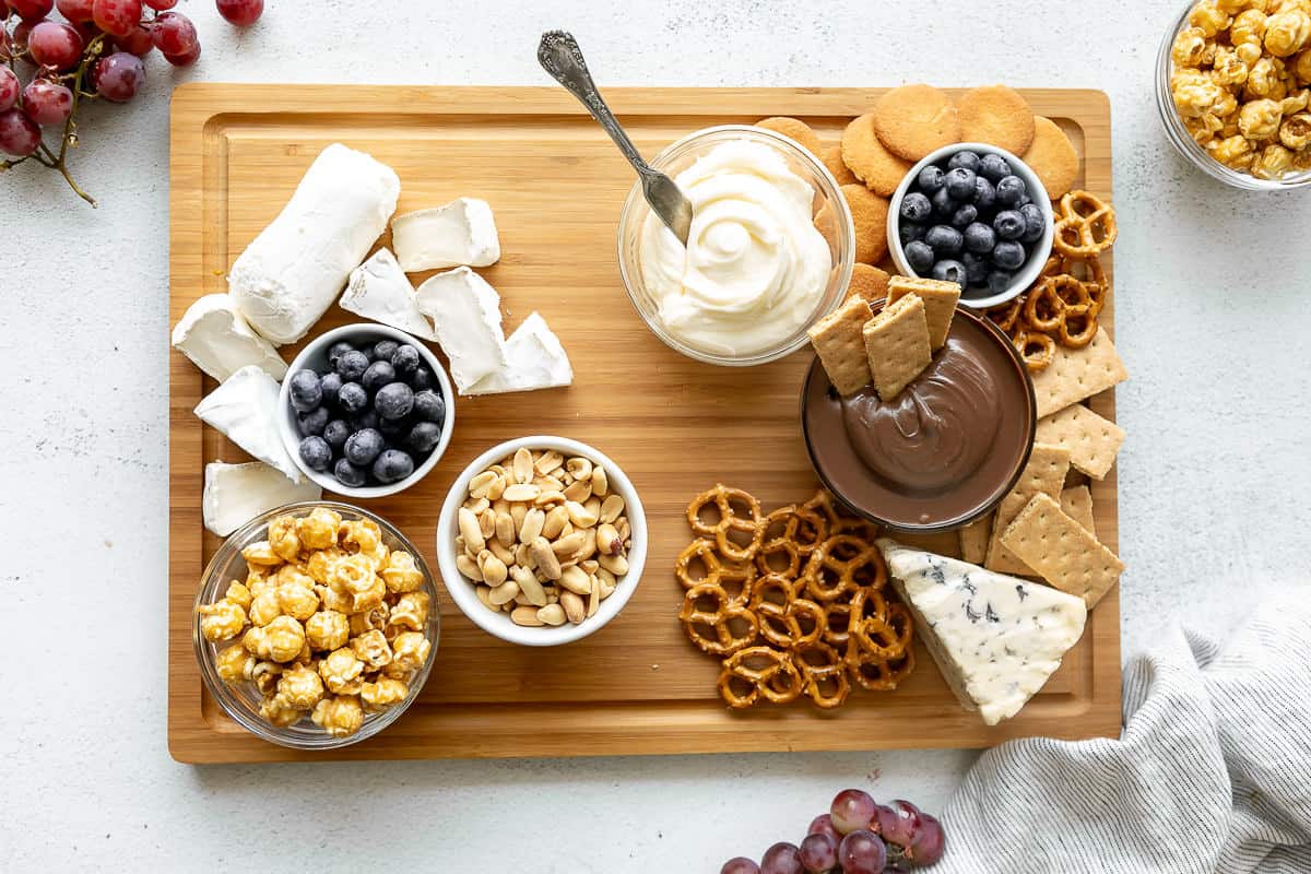 Dips, cheese, nuts, caramel corn, and cookies on a large cutting board.